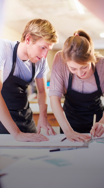 Close-up of two people wearing aprons and working together at a table. One is drawing something with a ruler.